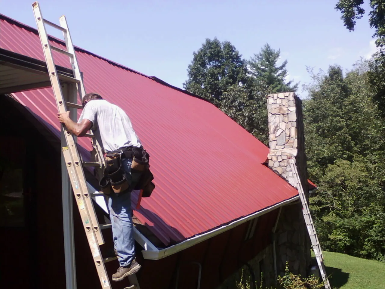 Roofer on a ladder beside a red metal roof and stone chimney — Soffit & Fascia Repair by Roofing Brothers in Sixes